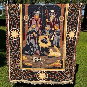 May include: A woven tapestry throw blanket featuring a nativity scene with figures in dark blue, gold, and maroon tones. The design includes a central manger, a lamb, and three figures standing above, all framed by a decorative border with star motifs.