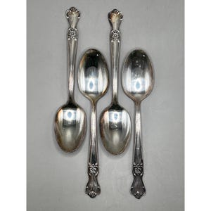 May include: Four silver-toned metal spoons arranged vertically. Two spoons feature ornate floral detailing at the top of the handle, while the other two feature similar floral detailing at the base of the handle. All spoons have a polished finish.