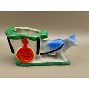Puede incluir: Una figura de cerámica de un buey blanco tirando de un carro. El buey tiene detalles azules en el lomo y la cabeza. El carro es blanco con una rueda naranja brillante, bordes negros y detalles verdes en el borde superior y la base.