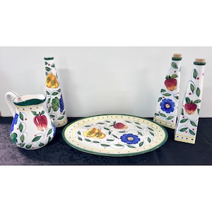 May include: A ceramic kitchen set featuring a pitcher, an oval platter, and three tall bottles with cork stoppers. Each piece is white with a hand-painted pattern of red apples, yellow pears, blue flowers, and green leaves with dark green trim.