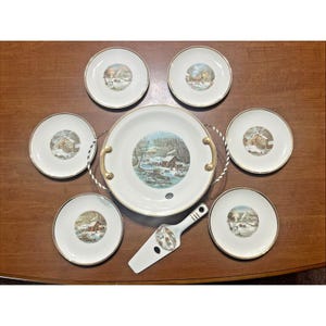 May include: A vintage serving set featuring a large round platter with gold handles, six small plates, and a cake server. Each piece has a winter scene design with a cabin in the snow and gold trim.