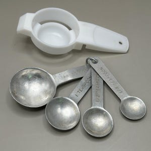 May include: A white plastic egg separator positioned above a set of four stainless steel measuring spoons held together by a metal ring. The spoons are labeled 1 tablespoon, 1 teaspoon, 1/2 teaspoon, and 1/4 teaspoon respectively.