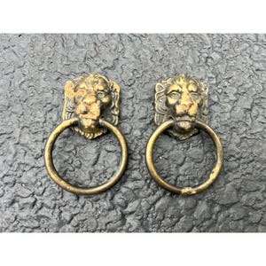 May include: Two antique brass lion head door knockers. Each knocker features a detailed lion's head with a circular ring handle. The brass has an aged, tarnished appearance, adding to their vintage charm. Perfect for adding a touch of classic elegance to any door.