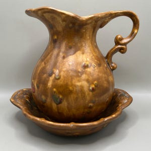 May include: A ceramic pitcher and matching basin set featuring a mottled brown glaze with raised, textured bumps and subtle hints of red and green. The pitcher has a curved handle and a scalloped rim, while the basin has a wide, fluted edge.