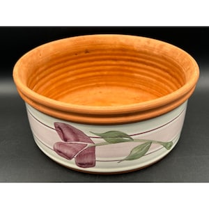 May include: A round ceramic bowl with a terracotta-colored interior and a white exterior. The outer surface features a horizontal light pink band decorated with a painted illustration of a dark purple tulip with green leaves and stems.