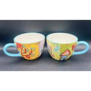 Può includere: Due tazze in ceramica con manici e bordi azzurri. L'esterno presenta un design dipinto a mano con accenti gialli, verdi e rossi, incluse illustrazioni di una casa e motivi floreali, con il testo parziale "LY" visibile.
