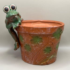 May include: A terracotta-colored ceramic planter featuring a sculpted green frog perched on the rim. The exterior of the round pot is decorated with embossed green clover leaf patterns and has a weathered, rustic finish throughout.