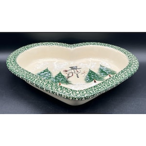 Puede incluir: Un molde de cerámica en forma de corazón con interior color crema que presenta un muñeco de nieve y pinos verdes pintados a mano. El borde tiene un patrón moteado en verde y blanco. El molde tiene un acabado liso y brillante en su totalidad.