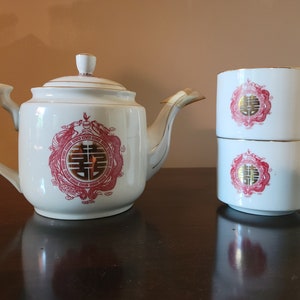 Puede incluir: Una tetera blanca y dos tazas de té blancas con caracteres chinos rojos y dorados. La tetera tiene un borde dorado y un diseño rojo y dorado con un símbolo de doble felicidad. Las tazas de té tienen un diseño rojo y dorado con un símbolo de doble felicidad.