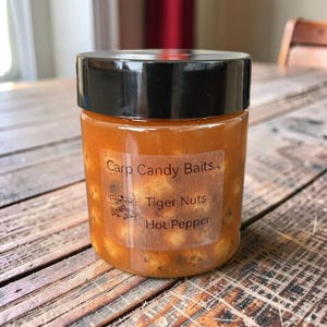 May include: A clear jar with a black lid filled with a reddish-orange liquid and small, round objects. The label reads "Carp Candy Baits, Tiger Nuts, Hot Pepper" with a fish logo. The jar sits on a wooden surface.