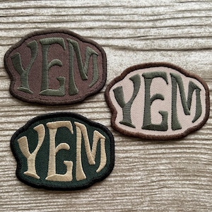 May include: Three embroidered patches with the letters "YEM" in a stylized font. The patches are in shades of brown, green, and beige, with a contrasting border. The patches are likely for clothing or accessories.