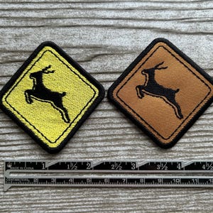 May include: Two embroidered patches featuring a black deer silhouette on a yellow and brown background. The patches are diamond-shaped with a black border. A ruler with inches and centimeters is at the bottom.