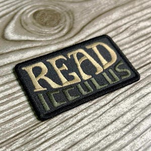 May include: A rectangular embroidered patch with a black background and a black stitched border. The word READ is embroidered in large, light tan letters, and the word ICCULUS is embroidered below in smaller, olive green letters.
