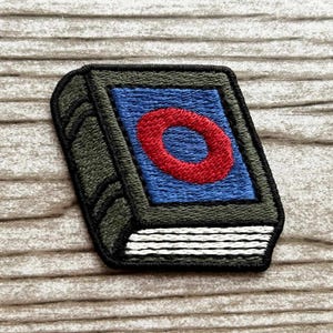May include: Embroidered patch in the shape of a book. The book has a blue cover with a red circle in the center. The edges are dark green with black stitching, and the pages are white.