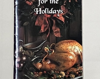 May include: A cookbook titled "Home for the Holidays" with a roasted turkey on the cover. The cover features a festive wreath, a red bow, and the text "Volume 5 - Holiday Recipes". The book is designed for holiday cooking.