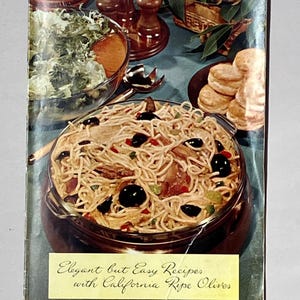 May include: A vintage cookbook cover featuring a glass casserole dish filled with spaghetti, black olives, and diced peppers. Beside it sits a bowl of green salad and a plate of biscuits. The text reads: Elegant but Easy Recipes with California Ripe Olives.