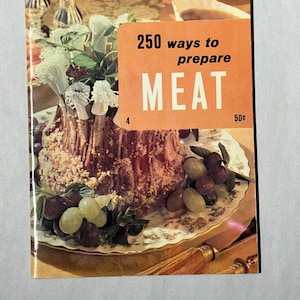 May include: A vintage cookbook titled "250 ways to prepare MEAT" with an orange cover. The book is placed on a decorative plate with a meat dish, grapes, and greenery. The cover also has the price of 50¢.