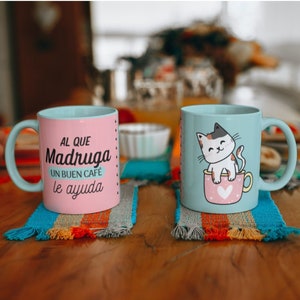 May include: Two ceramic mugs with different designs. One mug is pink with the text "Al que Madruga un buen cafe le ayuda" and the other mug is blue with a cartoon cat sitting in a pink mug with a heart.