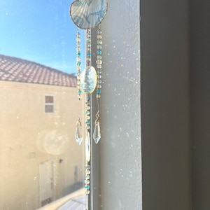 May include: A hanging suncatcher featuring a light blue and white seashell top with a small starfish accent. Three strands of clear and blue beads descend from the shell, interspersed with a central oval stone and clear crystal teardrop pendants.