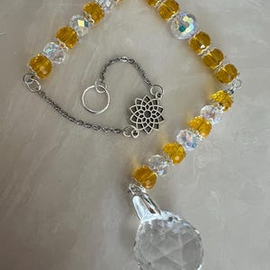 May include: A decorative suncatcher with yellow and clear crystal beads and a large faceted crystal pendant. The suncatcher has a silver chain and a decorative silver charm. The beads are strung together to form a triangle shape.
