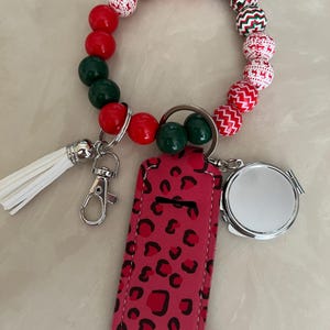 May include: A beaded wristlet keychain with red, green, and white beads. It includes a pink leopard print card holder, a small silver mirror, and a white tassel. The keychain has a silver clasp for easy attachment.