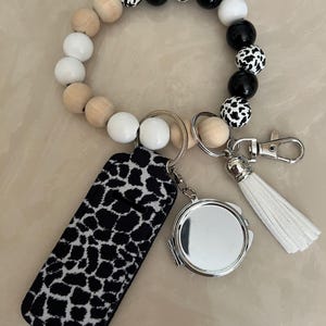 May include: A beaded keychain wristlet with a black and white cow print phone case, a small round silver compact mirror, and a white tassel. The wristlet features wooden, white, and black beads.