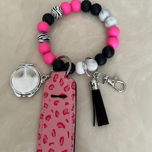 May include: A beaded keychain bracelet with a pink leopard print lip balm holder, a small round mirror, and a black tassel. The bracelet features pink, black, white, and zebra-striped beads, and a silver-colored clasp.