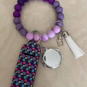 May include: A purple beaded wristlet keychain with a matching lip balm holder and a small silver compact mirror. The lip balm holder has a dark purple background with colorful polka dots. A white tassel hangs from the keychain.