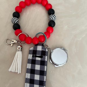 May include: A red, black, and checkered bead bracelet with a white tassel, a black and white plaid lip balm holder, and a small silver compact mirror. The bracelet is a wristlet style.