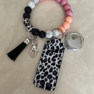 May include: A beaded keychain bracelet with a black tassel, a leopard print case, and a small round mirror. The bracelet features a mix of black, white, marble, pink, and peach-colored beads. Includes a silver clasp and key rings.