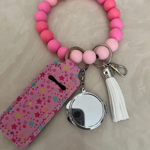 May include: A pink beaded bracelet with a keychain featuring a lip balm holder, a compact mirror, and a white tassel. The lip balm holder is pink with a star pattern.