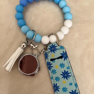 May include: A blue and white beaded bracelet with a white tassel, a round brown compact mirror, and a light blue lip balm holder with blue floral designs. The bracelet has a gradient of blue beads.