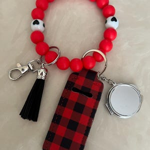 May include: Red beaded wristlet keychain with a black tassel, a red and black plaid lip balm holder, and a small round mirror. The wristlet includes white beads with black heart designs.