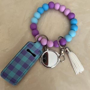 May include: A wristlet keychain with a gradient of blue, purple, and pink beads. It includes a lip balm holder with a blue and purple plaid pattern, a small round mirror, and a white tassel.