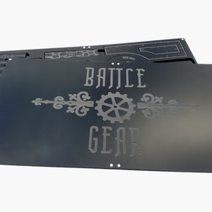 May include: Black metal game board with the words "BATTLE GEAR" in a decorative font. The board has a gear and flourish design. A second board is visible behind the first, with a grid and other cutouts.