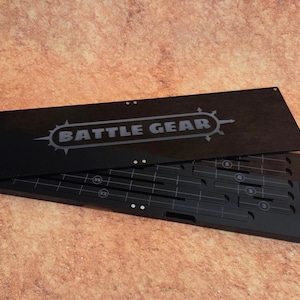May include: Black acrylic game organizer with the text "BATTLE GEAR" on the lid. The organizer is open, revealing compartments and markings for game pieces. The design is for tabletop gaming.