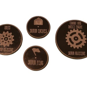 May include: Four round wooden tokens with brown and black coloring. Each token has a different symbol and text. The symbols include a gear, a flag, a chest, and a gear. The text includes "Units 50mm Objective", "Cohort and Battle Engine 50mm Objective", "30mm Caches", and "30mm Flag".