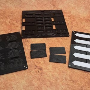 May include: Three black rectangular game boards with laser-cut slots and white arrow inserts. The boards feature text including "TURN 1", "TURN 2", "TURN 3", "TURN 4", "TURN 5", "TURN 6", "TURN 7", "TURN 8", "TURN 9", "TURN 10", and "BIG BATTLE TRACKER".