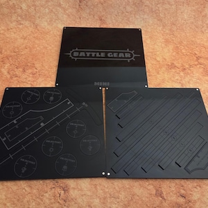 May include: Black acrylic gaming accessories with etched designs. The set includes a central piece with the text "BATTLE GEAR" and two hinged panels with various game templates and objective markers. The word "MINI" is also visible.
