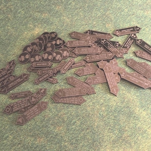 May include: A collection of dark gray game tokens, including circular pieces and various shapes resembling crosses and banners. The tokens are scattered on a green surface, with text visible on some of them.