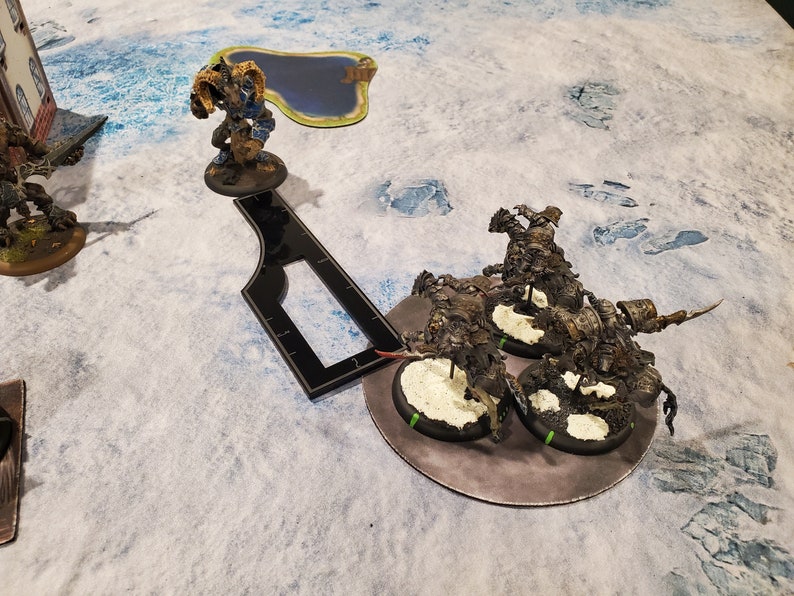 Puede incluir: Figuras en miniatura de personajes de fantas&iacute;a en un tablero de juego. Una figura es un guerrero azul y dorado con una bandera. Varias otras figuras son criaturas oscuras y blindadas. Una herramienta de medici&oacute;n negra est&aacute; presente.