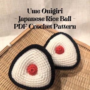 May include: Two crocheted white rice balls with black outlines and red centers, resembling Japanese onigiri, on a woven bamboo surface. The text "Ume Onigiri - Japanese Rice Ball - PDF Crochet Pattern" is above the rice balls.