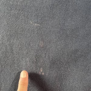 May include: Close-up of a dark navy blue fabric with a textured surface. The fabric has several small, light-colored spots and a few pinkish stains. A finger is visible in the lower portion of the image.