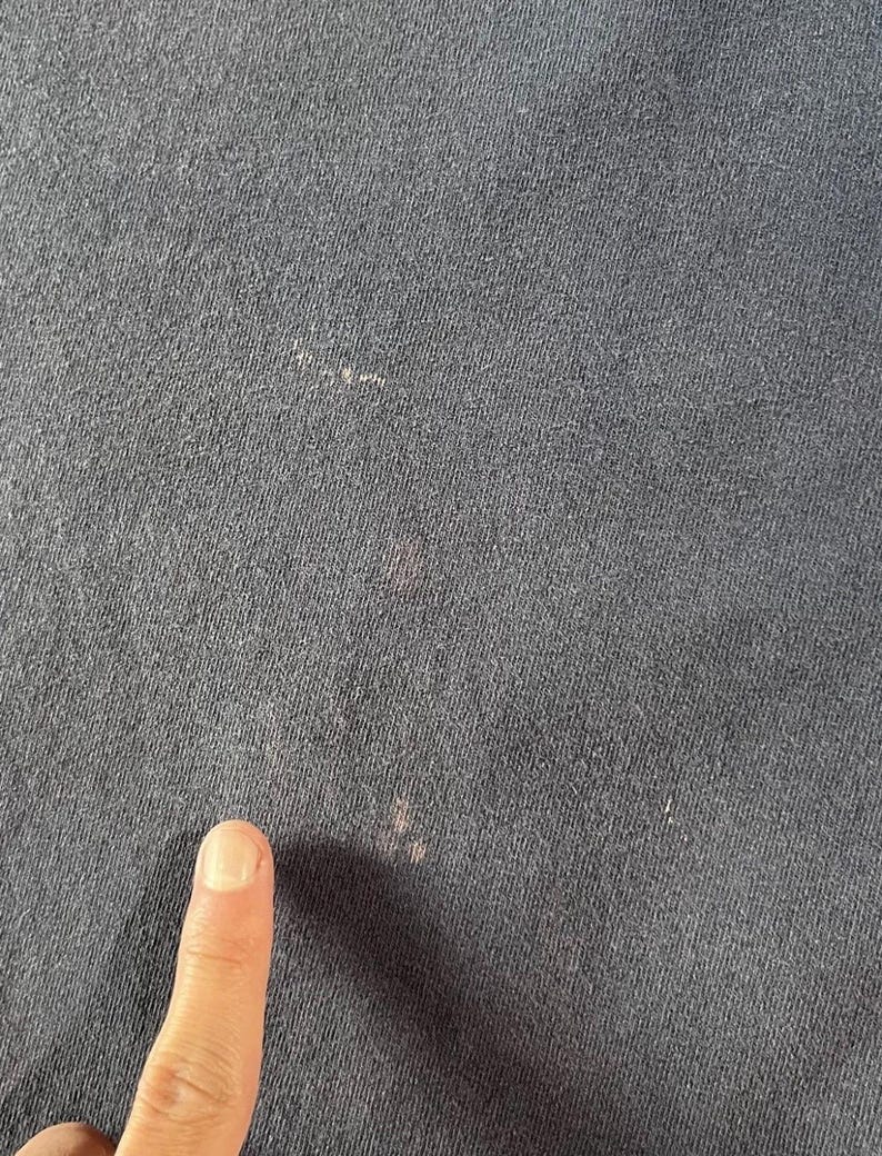 May include: Close-up of a dark navy blue fabric with a textured surface. The fabric has several small, light-colored spots and a few pinkish stains. A finger is visible in the lower portion of the image.