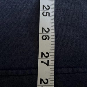 May include: A navy blue fabric is measured with a white and black measuring tape. The tape shows measurements from 24 to 28 inches. The fabric appears to be a t-shirt or similar garment.