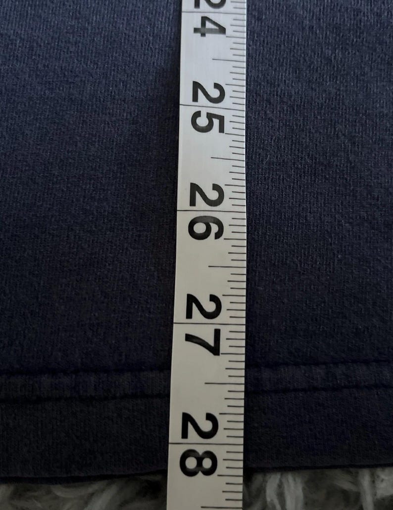 May include: A navy blue fabric is measured with a white and black measuring tape. The tape shows measurements from 24 to 28 inches. The fabric appears to be a t-shirt or similar garment.