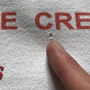 Pu&ograve; includere: Un capo in tessuto grigio chiaro con testo stampato in rosso che recita "ACE CREW" e "phs". &Egrave; presente un piccolo foro visibile nel tessuto situato direttamente sotto la parola "ACE" e sopra il dito che indica l'area danneggiata.