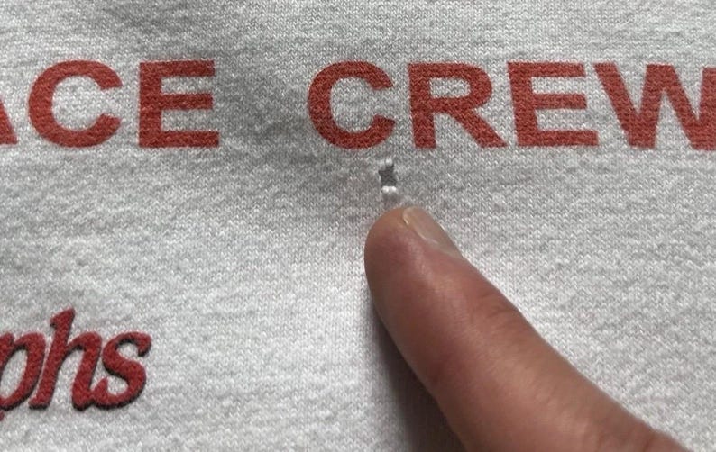 Pu&ograve; includere: Un capo in tessuto grigio chiaro con testo stampato in rosso che recita "ACE CREW" e "phs". &Egrave; presente un piccolo foro visibile nel tessuto situato direttamente sotto la parola "ACE" e sopra il dito che indica l'area danneggiata.