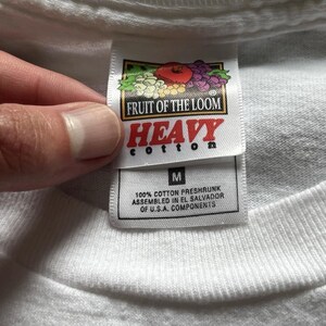 Pu&ograve; includere: Una t-shirt bianca con un'etichetta del marchio Fruit of the Loom Heavy Cotton. L'etichetta mostra il logo del marchio, la parola HEAVY, la taglia M e il testo che indica 100% cotone prelavato, assemblato in El Salvador con componenti statunitensi.
