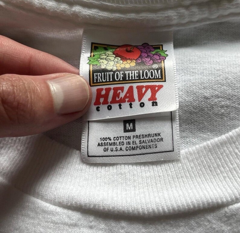 Pu&ograve; includere: Una t-shirt bianca con un'etichetta del marchio Fruit of the Loom Heavy Cotton. L'etichetta mostra il logo del marchio, la parola HEAVY, la taglia M e il testo che indica 100% cotone prelavato, assemblato in El Salvador con componenti statunitensi.